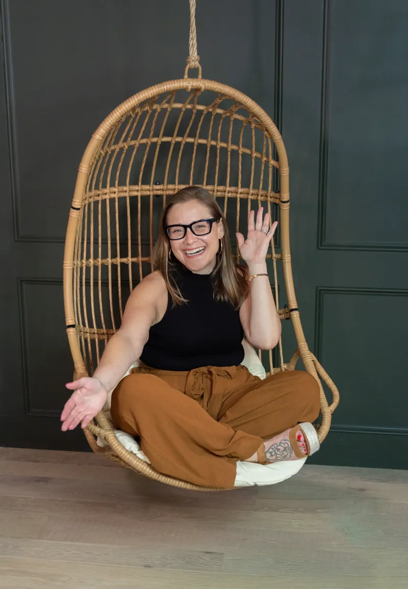 Maria Englehardt, founder of The Inkwell Suite, smiling and waving while seated in a hanging chair